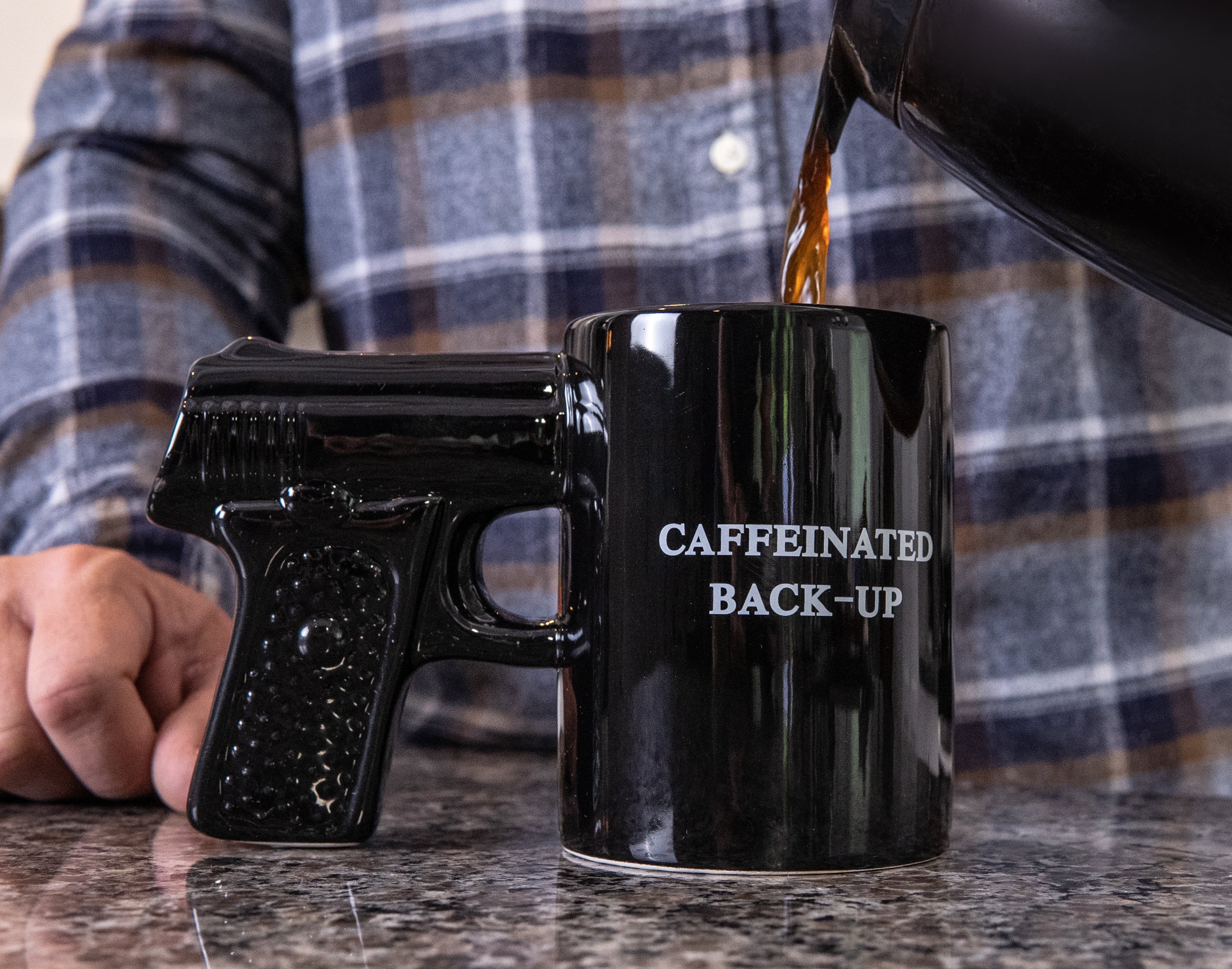 Black mug with 'CAFFEINATED BACK-UP' text being filled with coffee, held by a person wearing a plaid shirt.