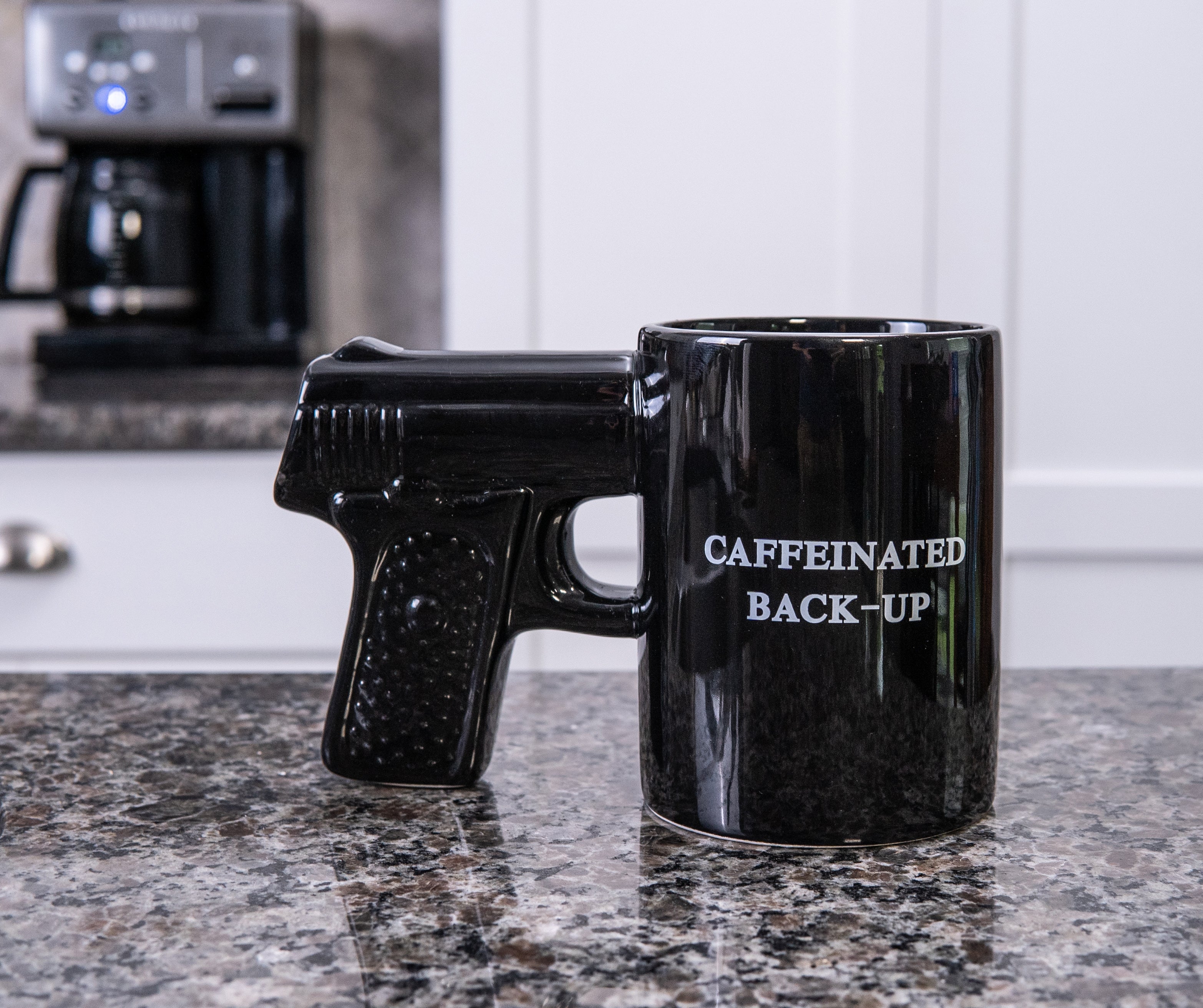 Black mug shaped like a gun with 'CAFFEINATED BACK-UP' text on a kitchen counter.