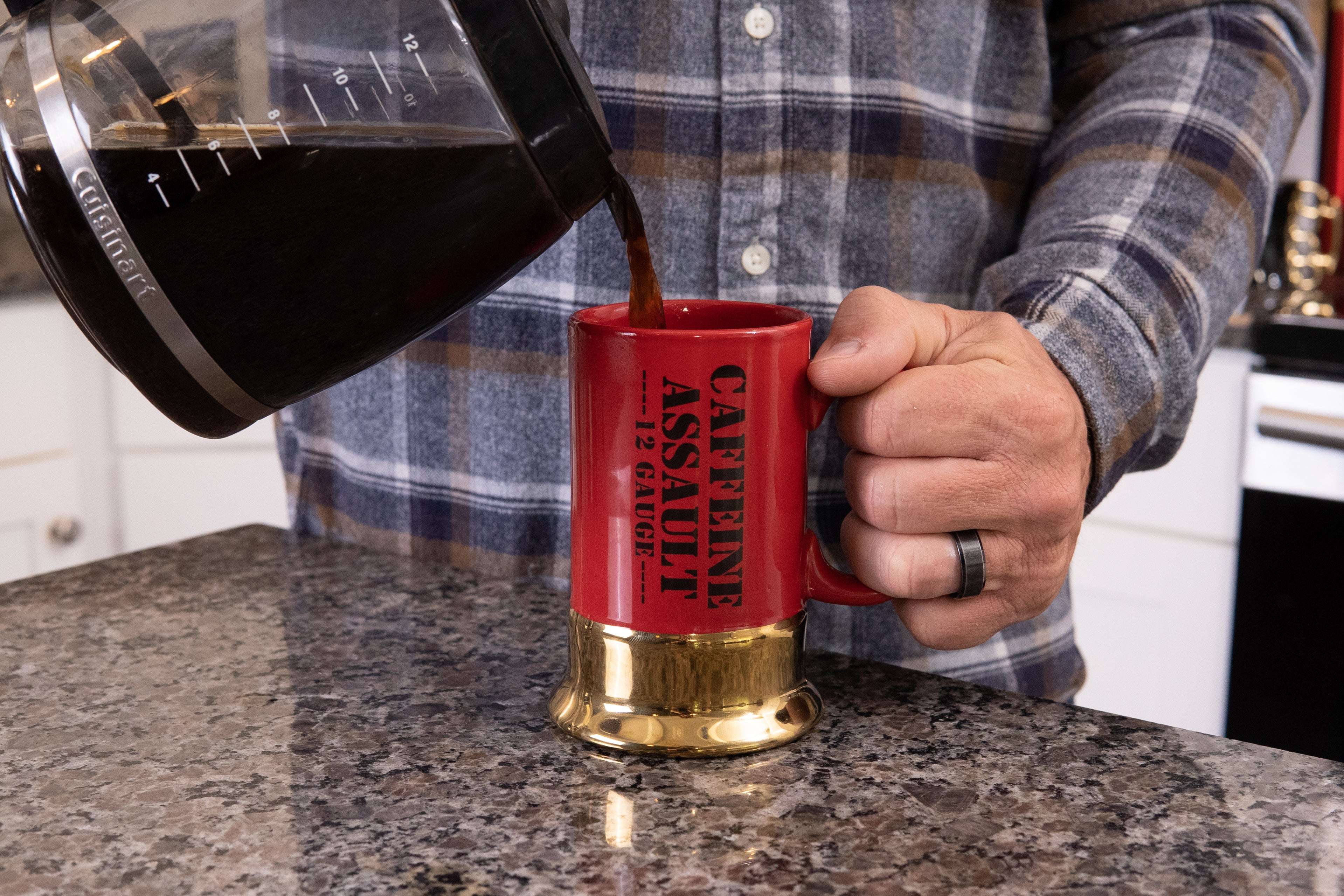 Person pouring coffee from a carafe into a red mug with gold base, set against a kitchen counter.