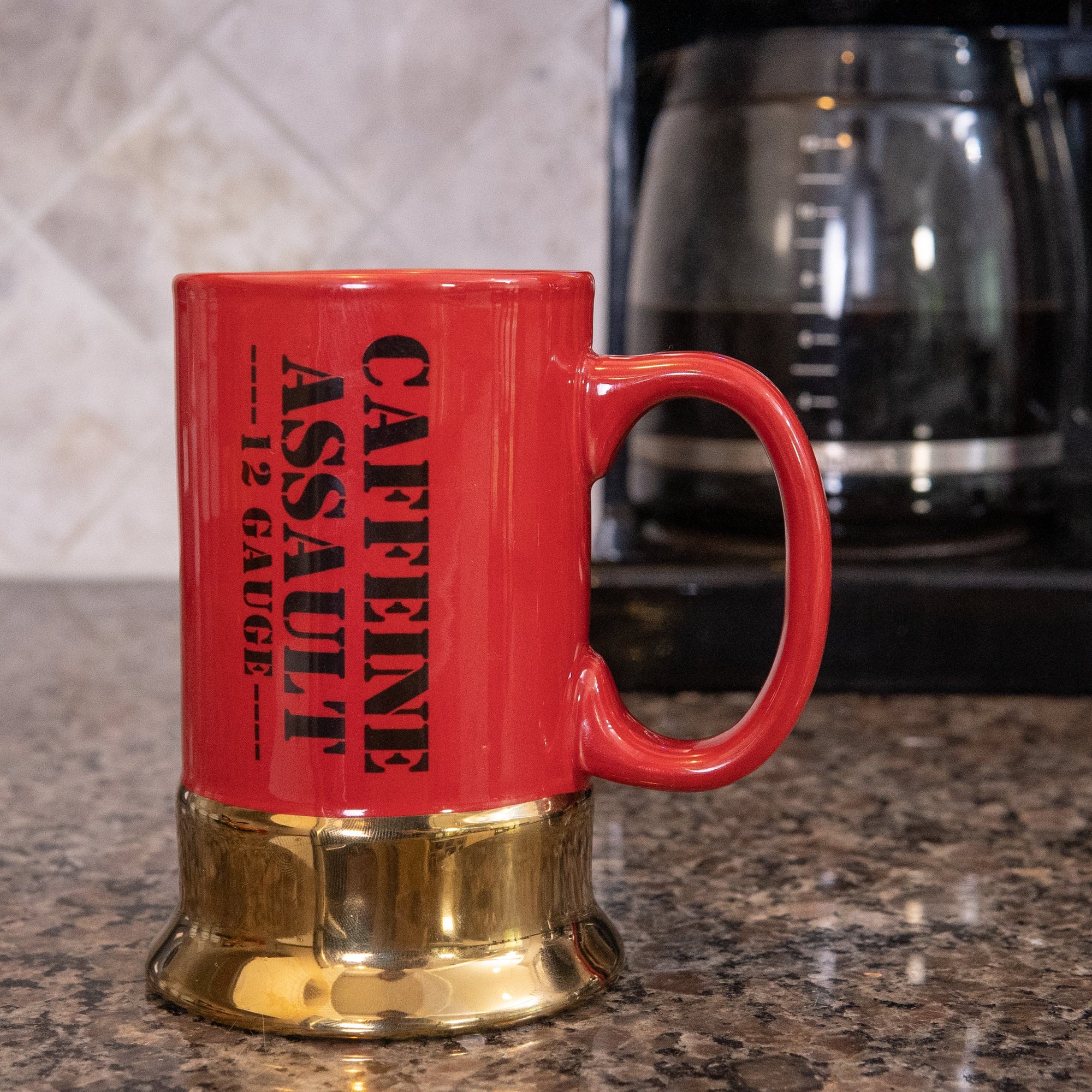 Red mug with 'Caffeine Assault' text on a kitchen counter with a coffee maker in the background.