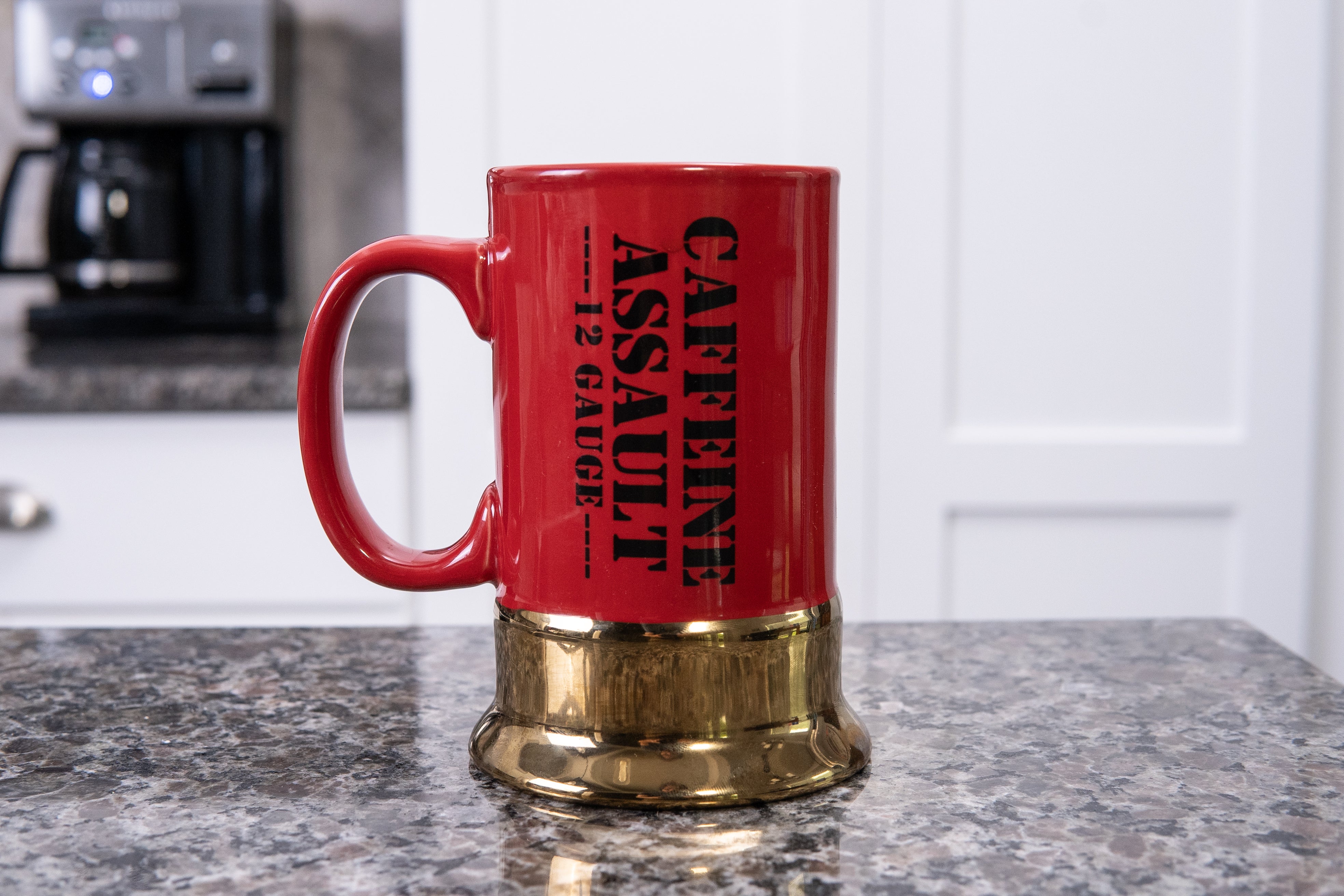Red mug with gold base on a kitchen counter, featuring text 'CAFFEINE ASSAULT 12 GAUGE'.