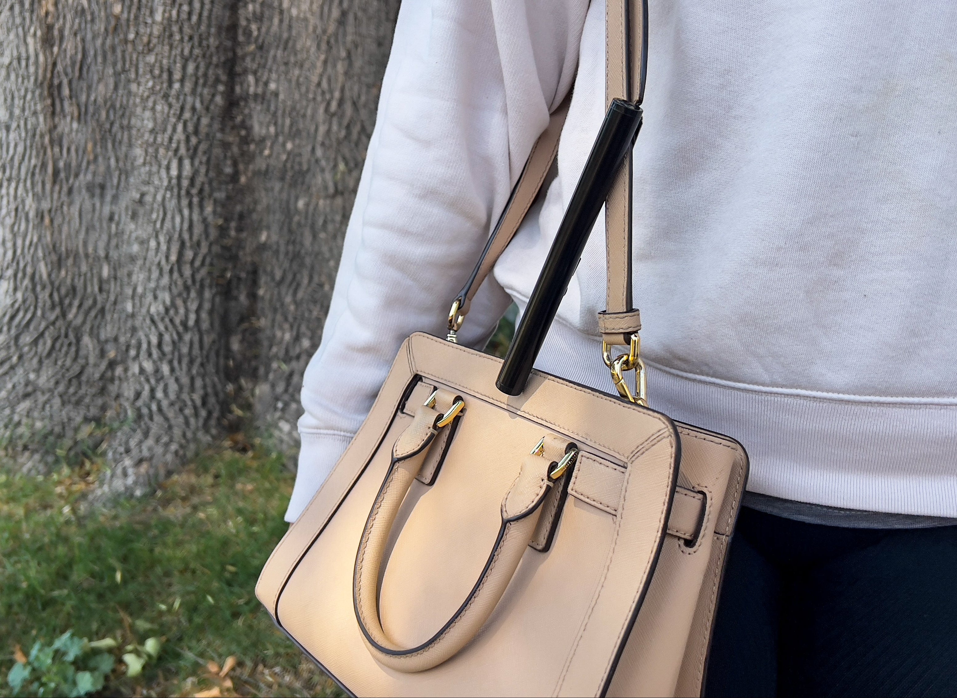 Beige handbag held by a person wearing a light gray sweatshirt, with a tree trunk in the background.