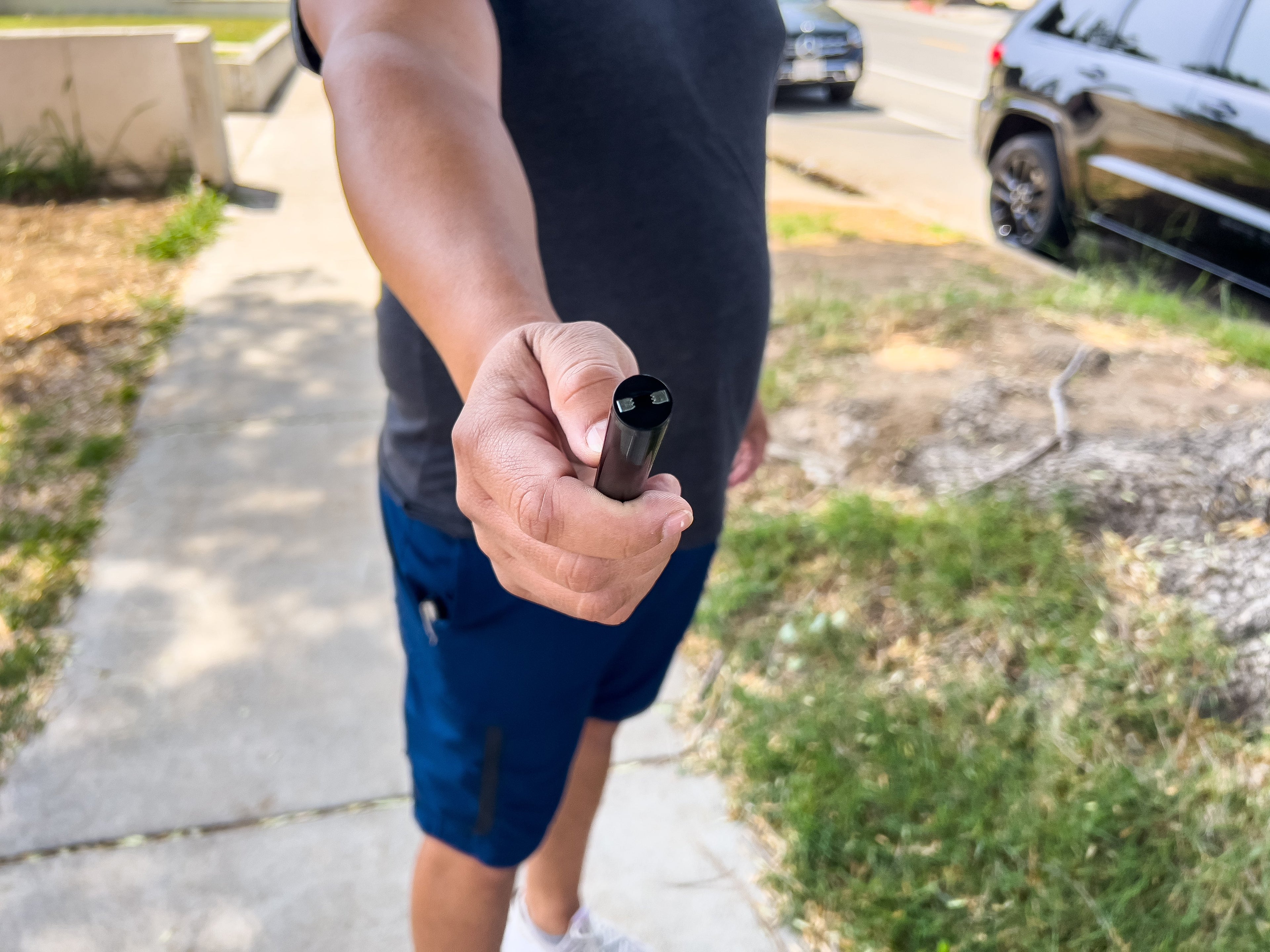 Person holding a black cylindrical object on a sidewalk with cars in the background