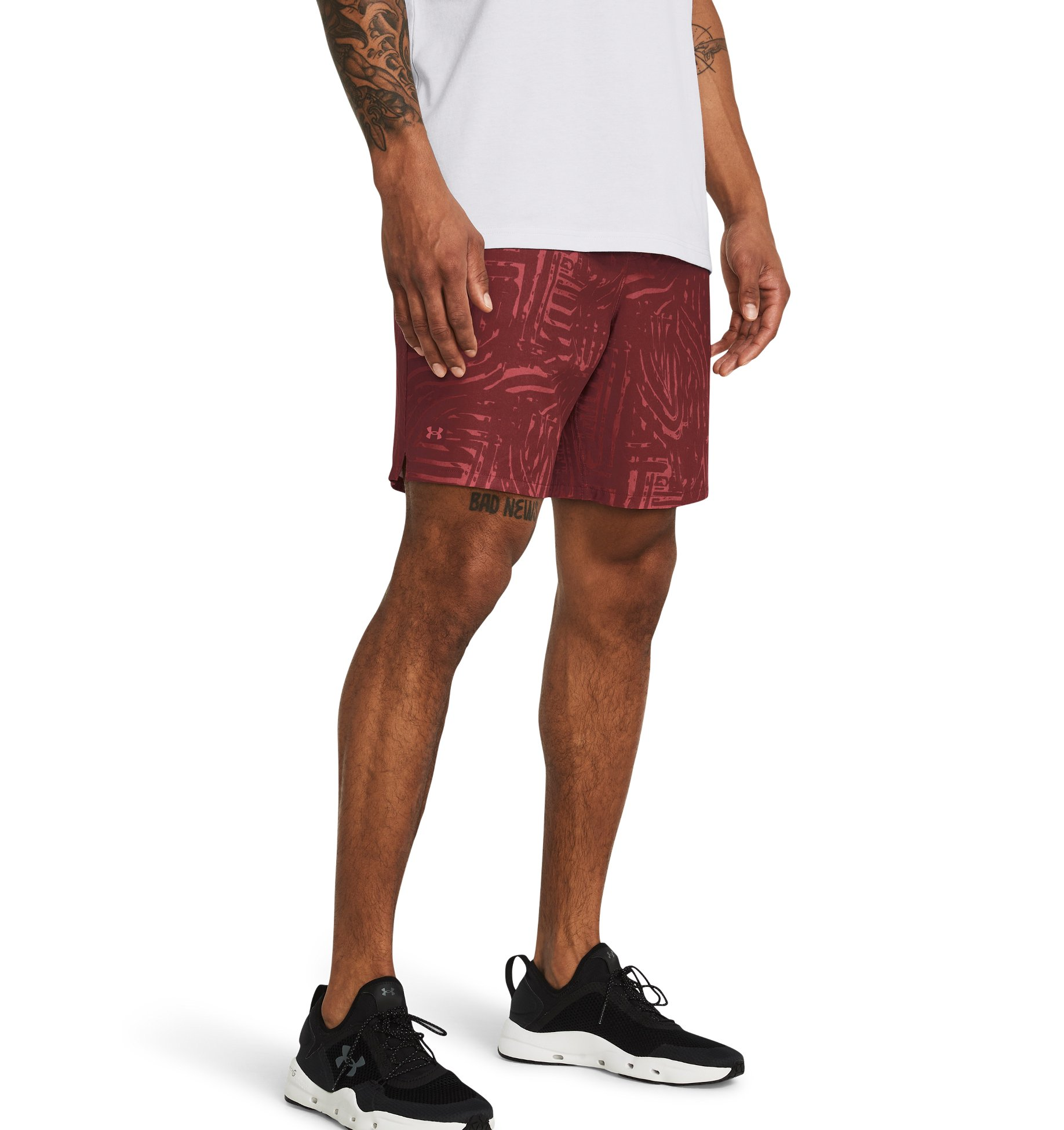 A man wearing Under Armour men's UA Expanse 2-in-1 boardshorts in a red and black pattern. He is also wearing a white T-shirt and black sneakers.