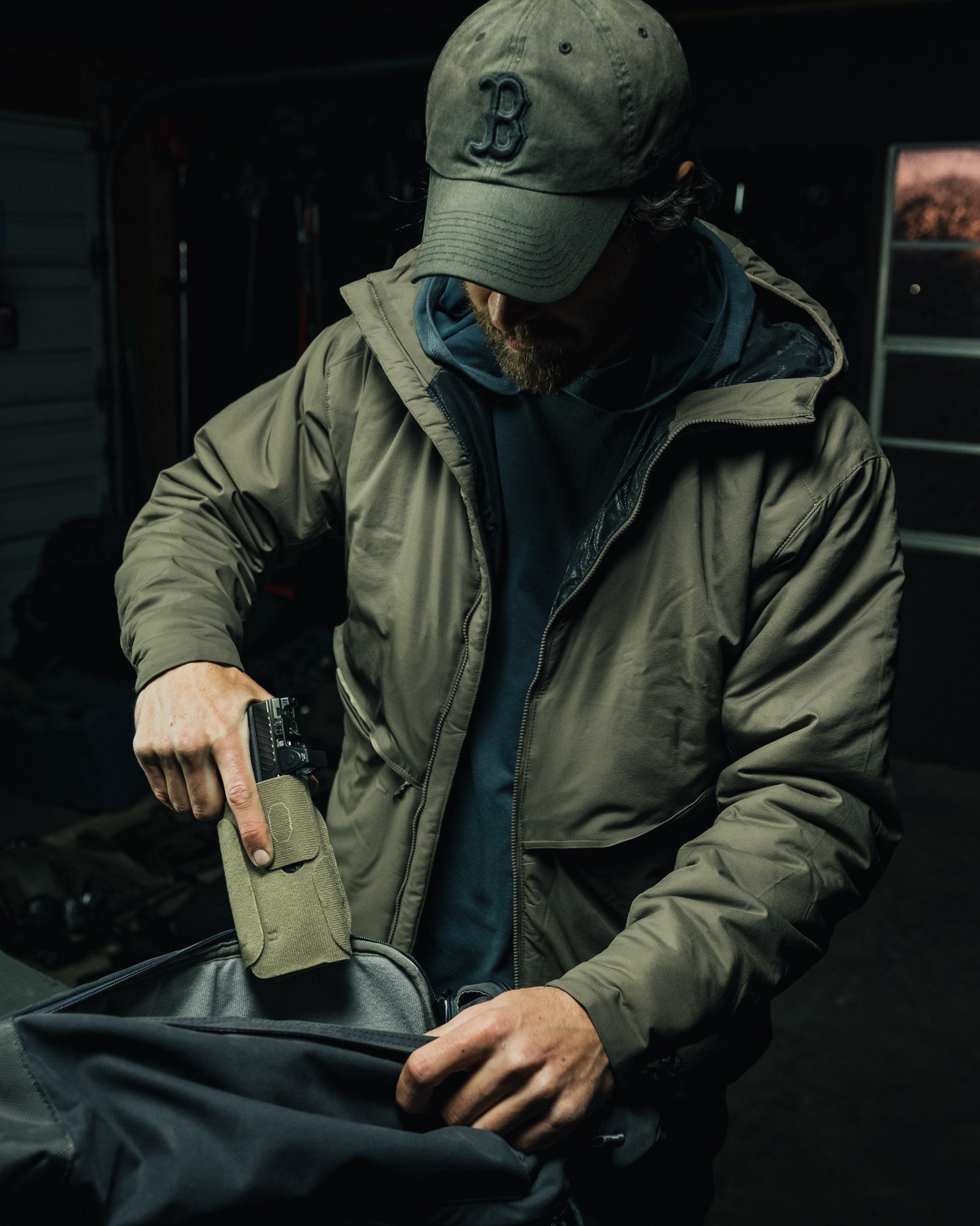 Person in a green jacket and cap holding a phone and a pouch, sitting in a dark setting.