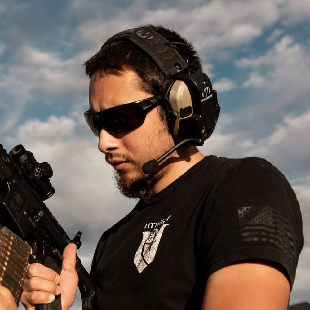 Man wearing sunglasses and a headset, holding a rifle against a cloudy sky.