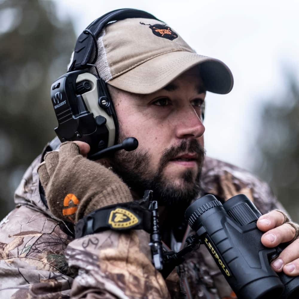 Hunter wearing camouflage gear, Walker's Razor Walkie Talkie Attachment GWP-RZRWT, and binoculars in a forest setting