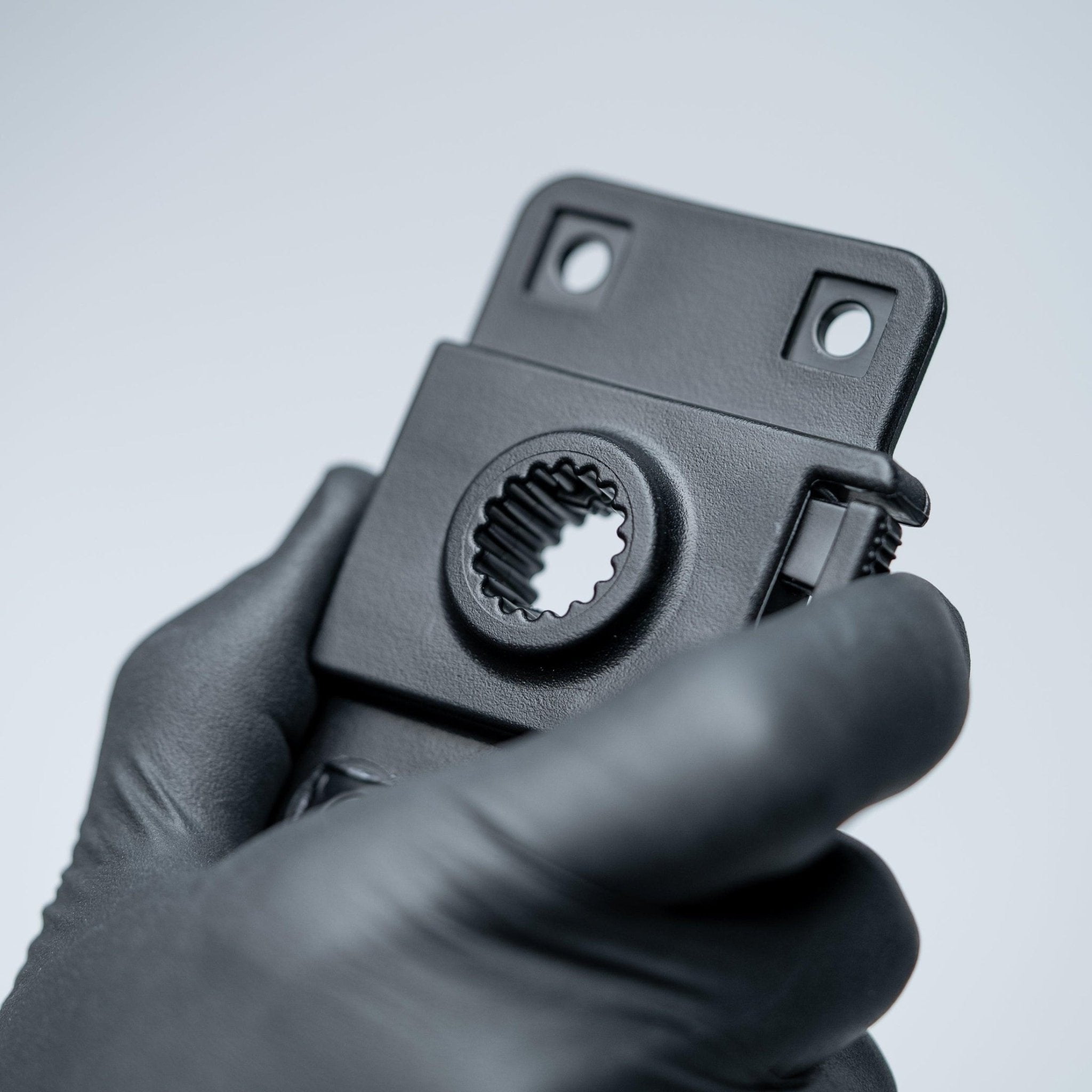 A close-up of a black plastic mounting component held by a hand wearing a black glove, set against a clean light gray background.