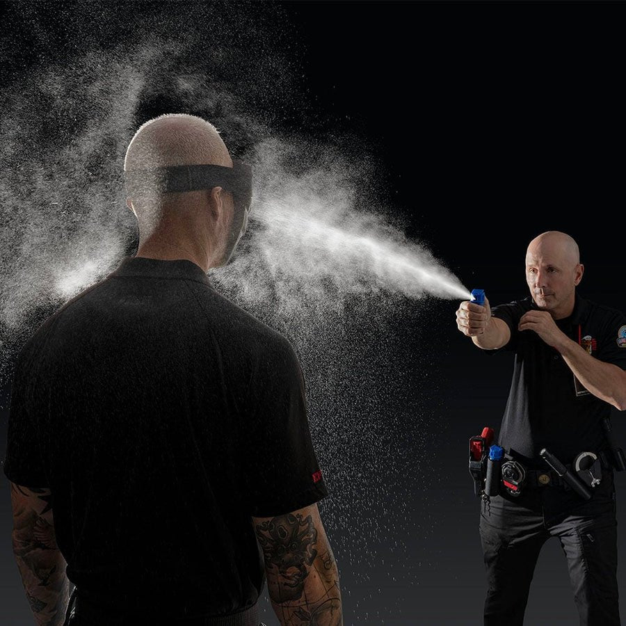 Officer sprays a blue-top ASP Defender inert fog canister at a role player wearing eye protection during OC training