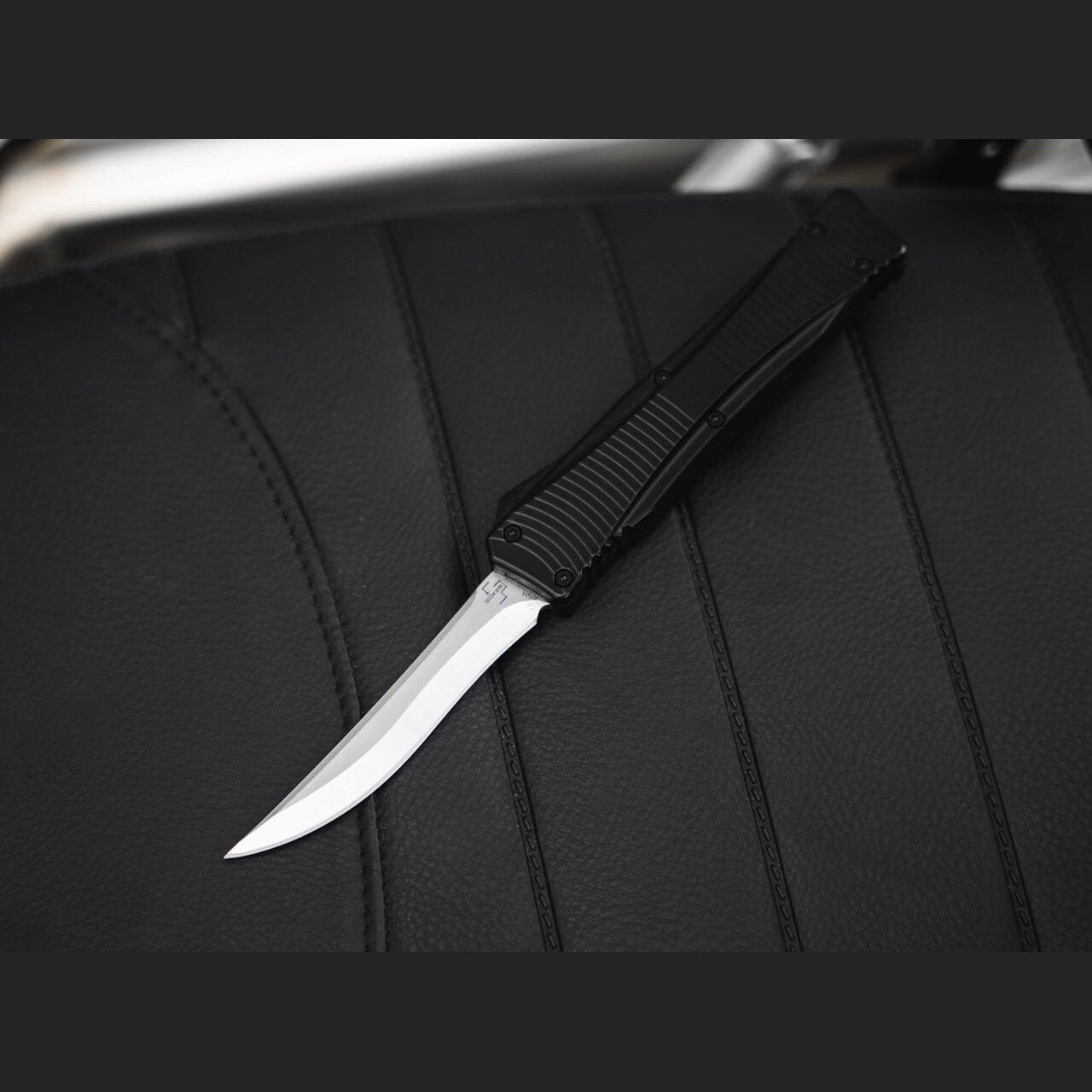 Böker Plus Eagle 2.0 OTF automatic knife with recurve blade deployed on a black leather seat