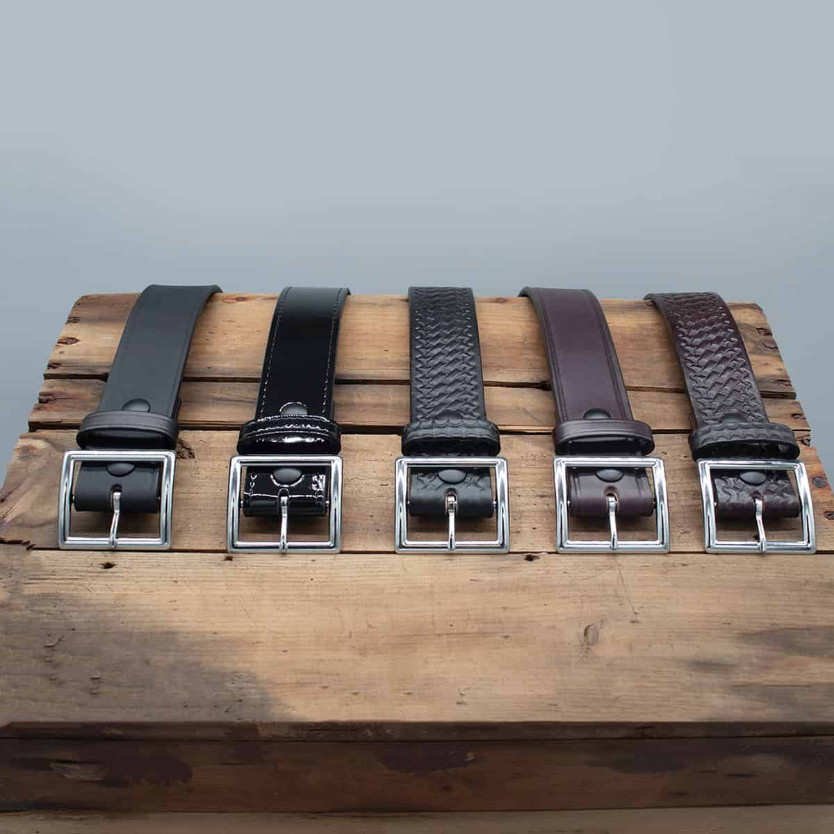 Four leather belts with silver-toned buckles displayed side by side on a rustic wooden block against a plain gray background.