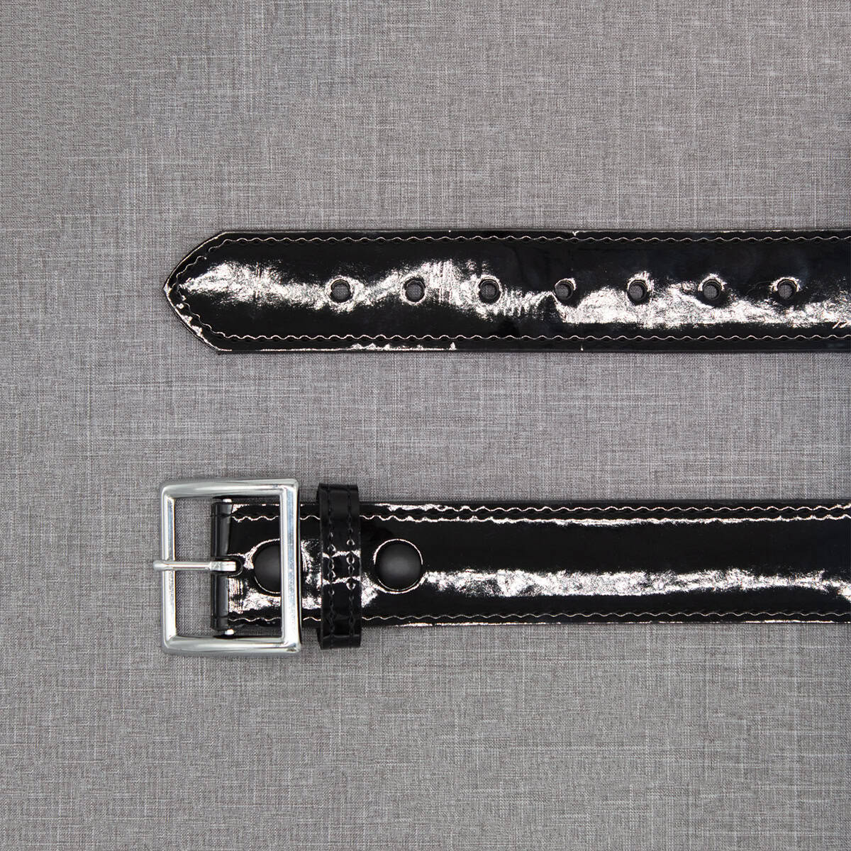 A glossy black patent leather watch strap with a silver-tone metal buckle, laid flat on a textured gray fabric background.