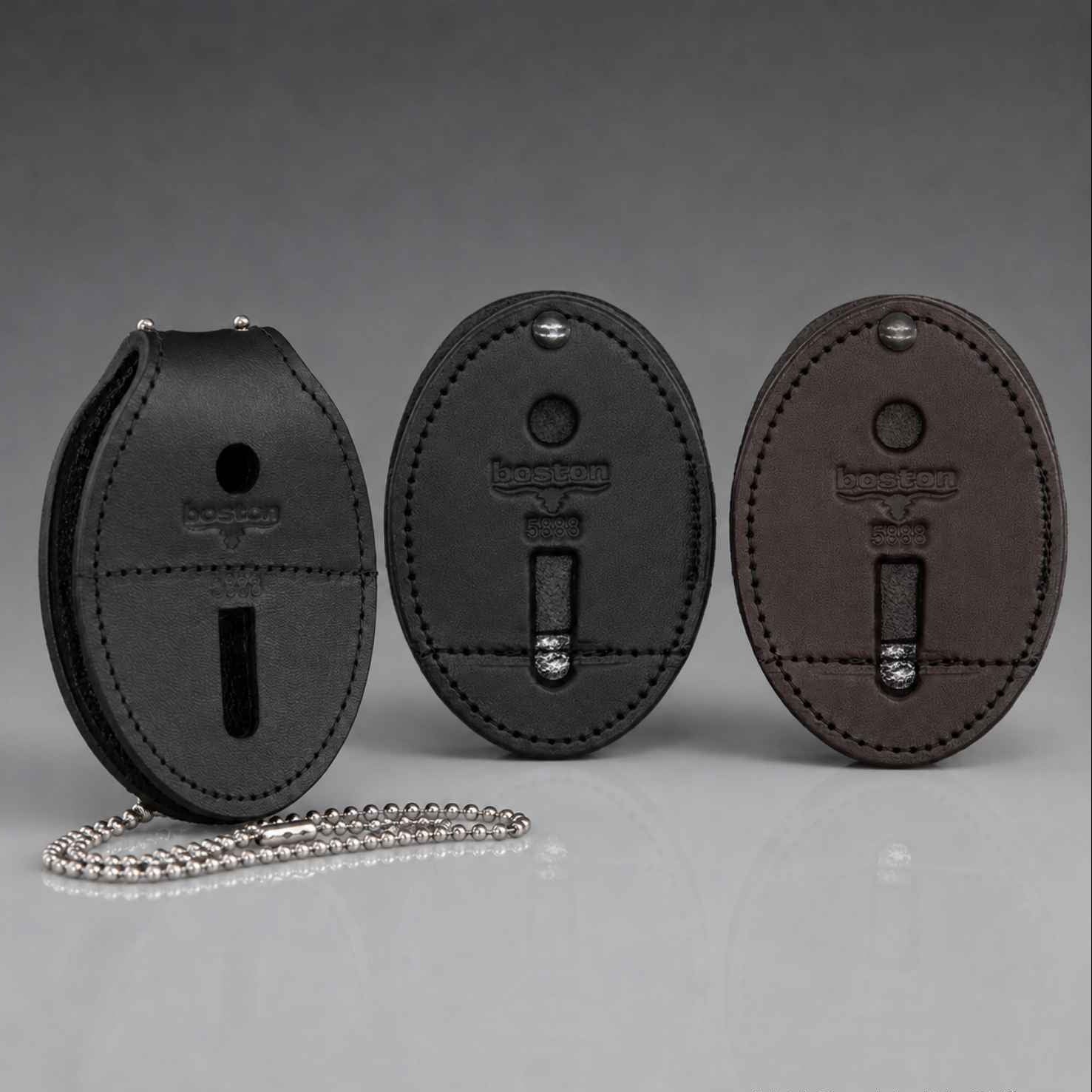 Three oval leather badge clips in black, dark gray, and brown, each with a silver ball chain, are displayed on a reflective gray surface against a gradient gray background.