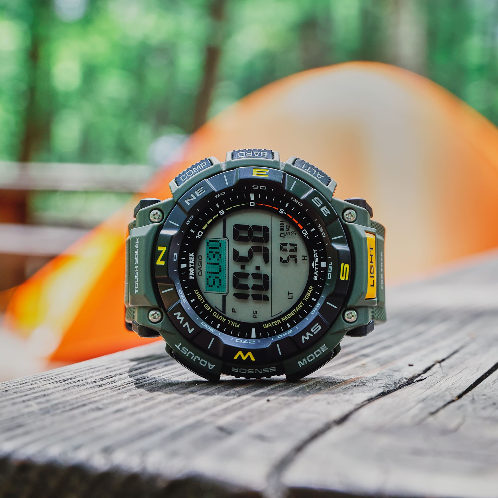 Digital watch on a wooden surface with a blurred camping scene in the background