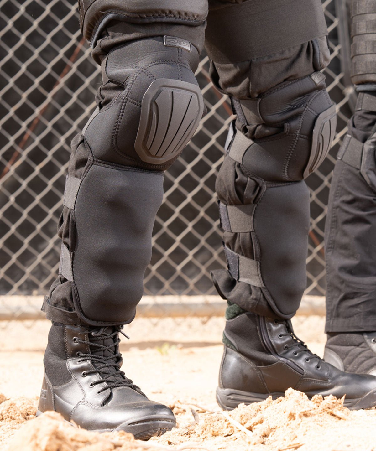 Close-up of protective knee pads and boots worn by a person on a chain-link fence background.
