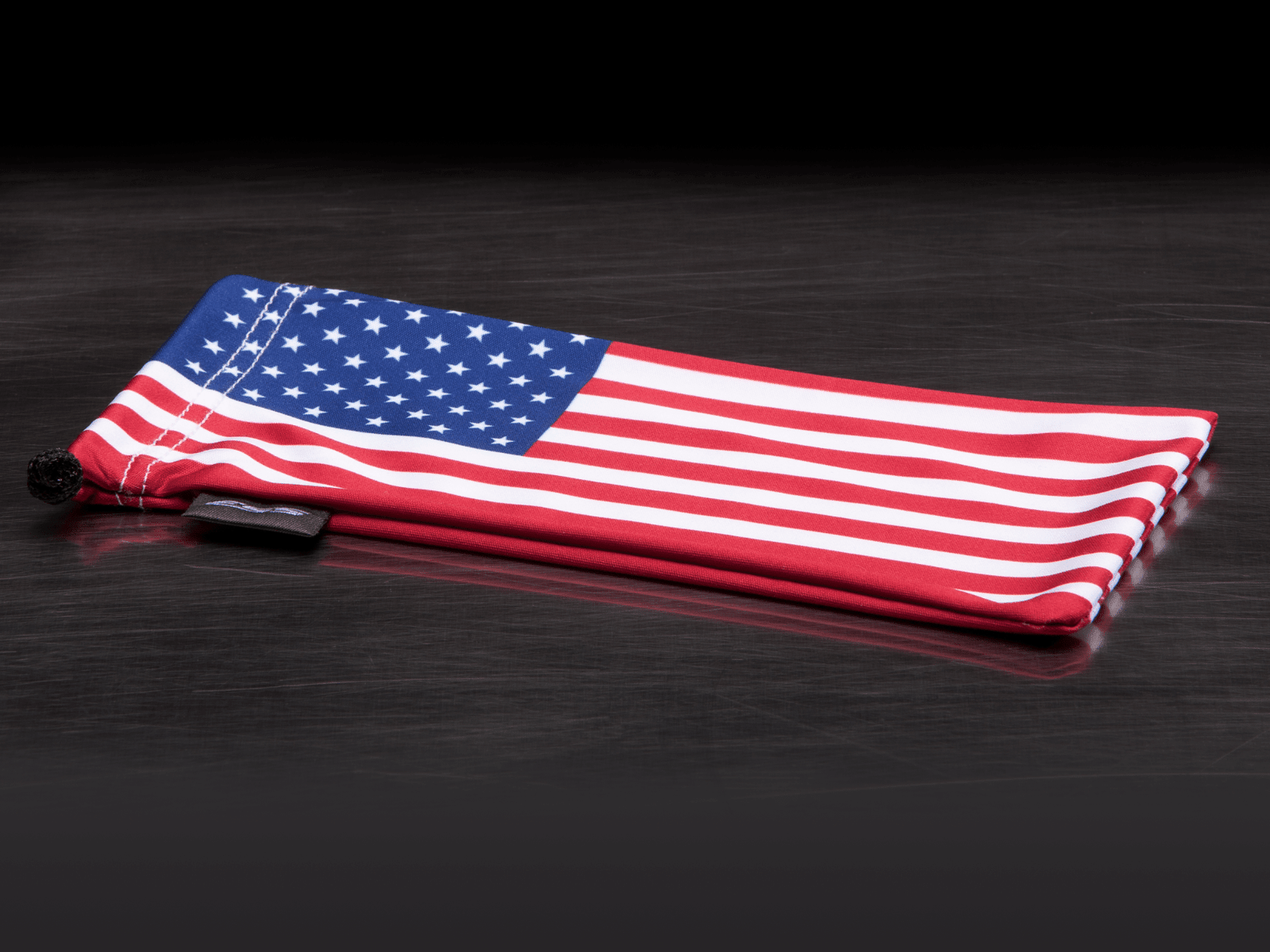 A folded fabric pouch featuring the American flag pattern with red and white stripes and a blue field of white stars, shown on a dark reflective surface.