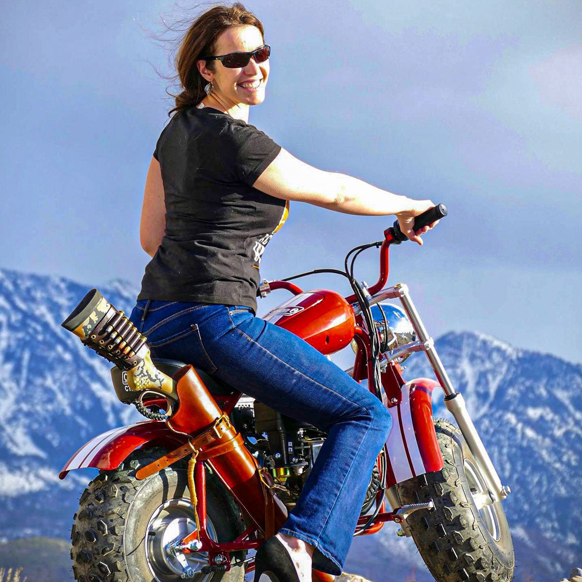 A woman wearing sunglasses, a black t-shirt, and blue jeans rides a red mini chopper motorcycle against a backdrop of snow-capped mountains and a blue sky.