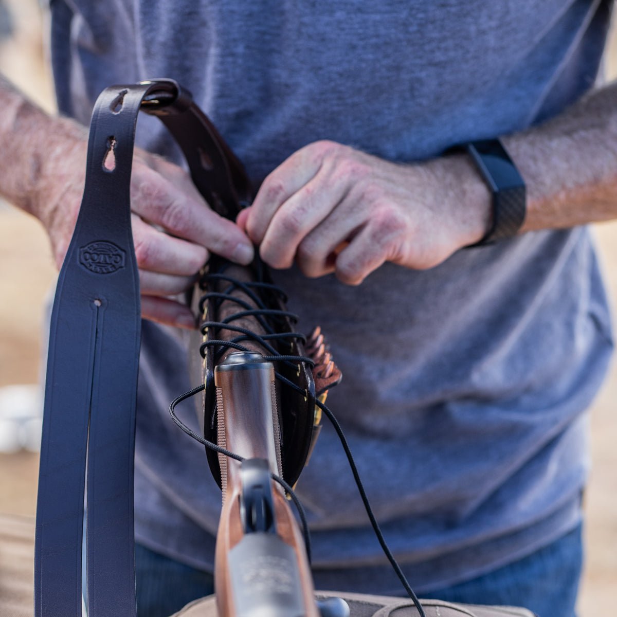 A close-up of a person's hands adjusting a black leather rifle sling on a shotgun. The person is wearing a blue-gray t-shirt and a black smartwatch.