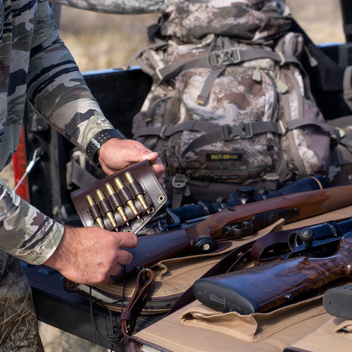 A person in a camouflage long-sleeve shirt holds an open leather case displaying brass-cased ammunition cartridges over a black truck bed. A large camouflage backpack and several rifles are arranged in the truck bed behind.