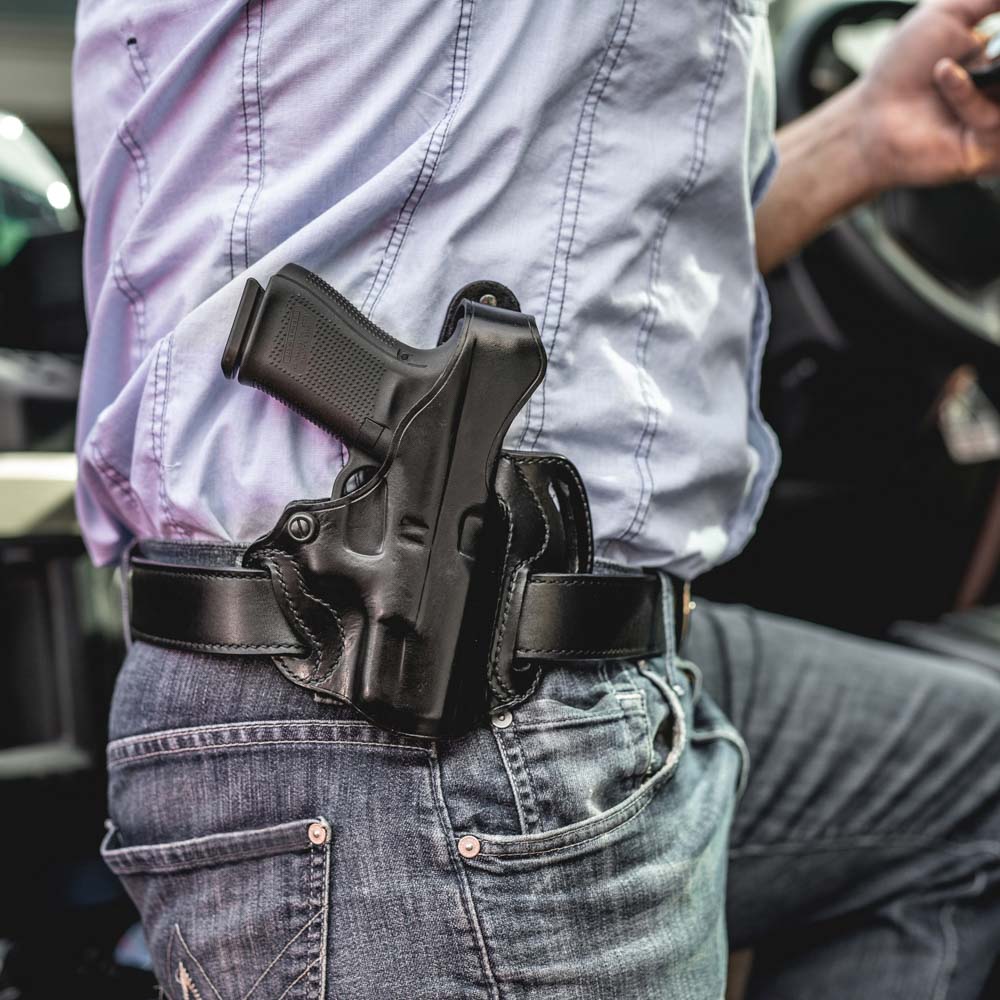 A black leather holster containing a handgun, worn at the waist on a person's hip over a light blue shirt and blue jeans.