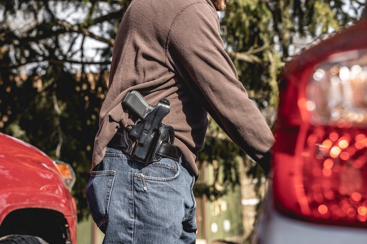 A black semi-automatic handgun in a black holster worn on a person's hip, shown outdoors between blurred red cars.