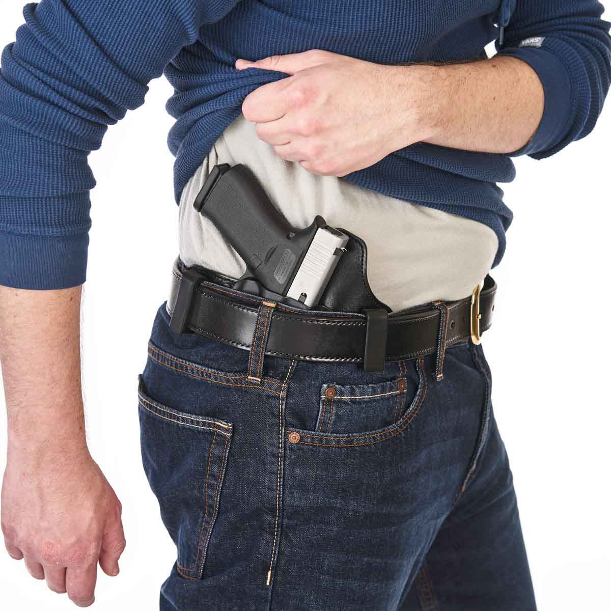 A close-up of a person wearing a black hip holster with a handgun inserted, over a light gray shirt and dark blue jeans, against a white background.