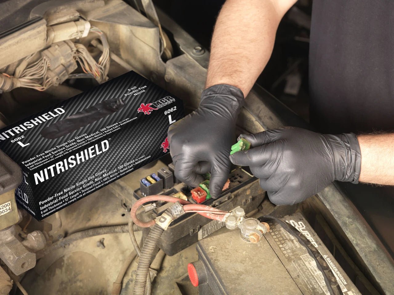 A person wearing black gloves handles a green electrical connector near a car battery, with a black Nitrishield box placed nearby in the engine bay.