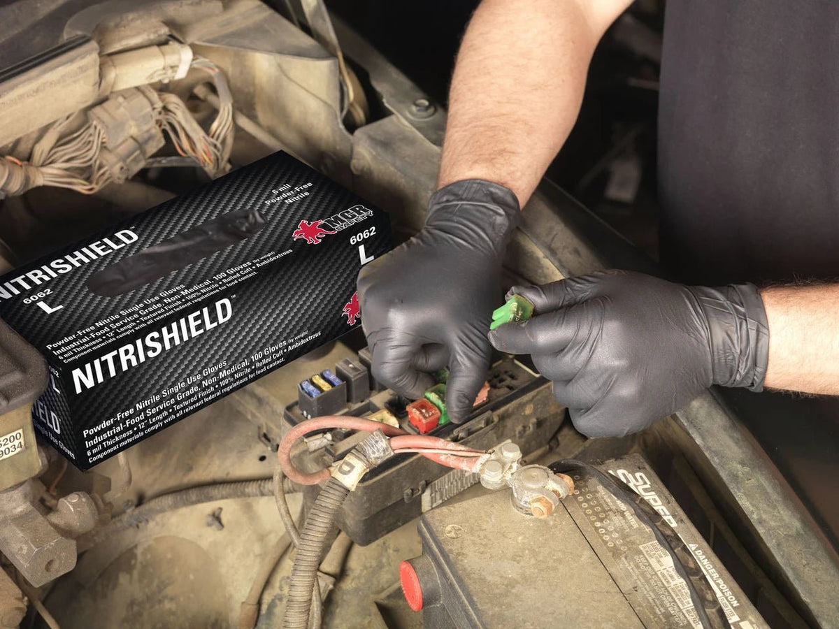 A person wearing black gloves handles a green electrical connector near a car battery, with a black Nitrishield box placed nearby in the engine bay.