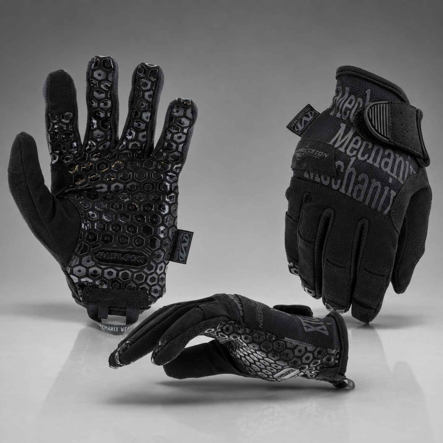 Three black Mechanix gloves shown from different angles on a reflective gray studio surface, displaying the palm, back of hand, and wrist closure.