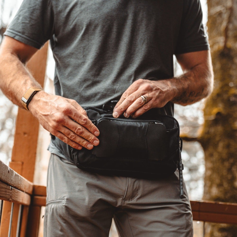 Person holding a black waist bag outdoors with wooden furniture and blurred natural background
