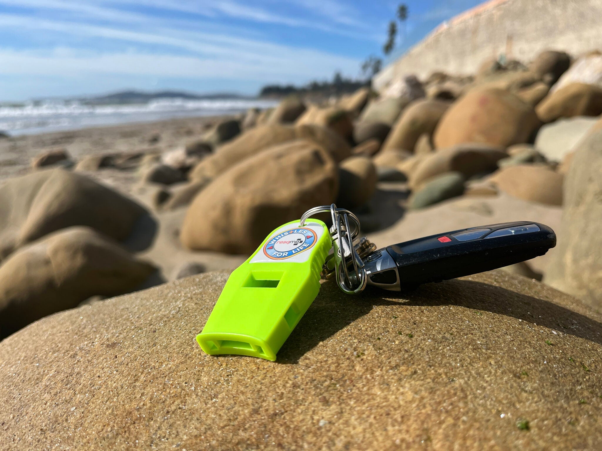 Green whistle and black car key on a rocky beach with ocean in the background