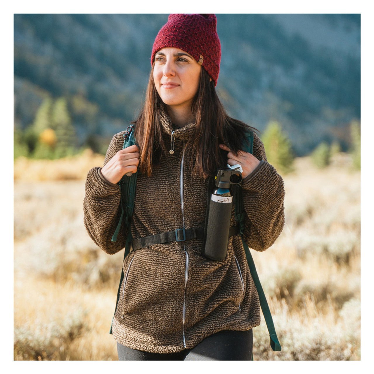 Woman in a brown jacket and red beanie holding a backpack and water bottle in a mountainous landscape.