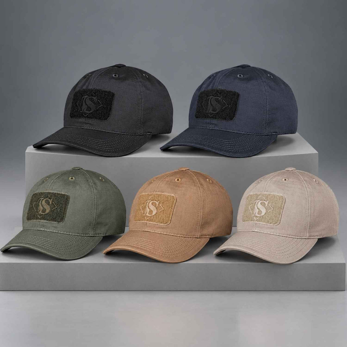 Four baseball caps in dark gray, navy, olive green, and beige/tan are arranged on white display blocks against a neutral gray studio background. Each cap has a rectangular embroidered patch on the front with a stylized letter S.
